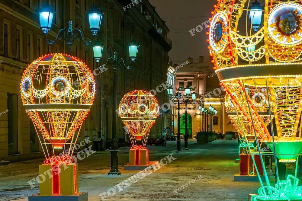 На фотографии изображена пешеходная улица в Санкт-Петербурге, украшенная к Новому году световыми инсталляциями в форме воздушных шаров. Яркие гирлянды и праздничная иллюминация придают улице волшебный вид. Старинные фонари освещают пространство, создавая теплую атмосферу в холодную зимнюю ночь. Этот праздничный декор делает город особенно привлекательным для прогулок во время зимних праздников.